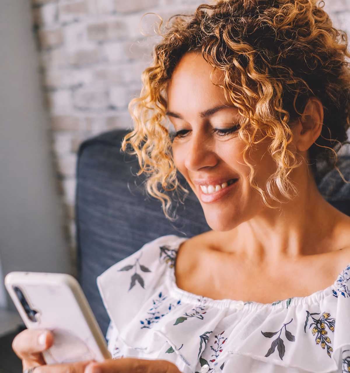 Smiling woman with curly hair looking at her smartphone while sitting on a gray couch.