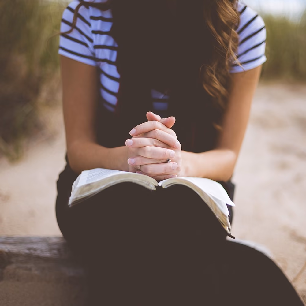 Woman with clasped hands praying over an open Bible, seated outdoors on a beach.
