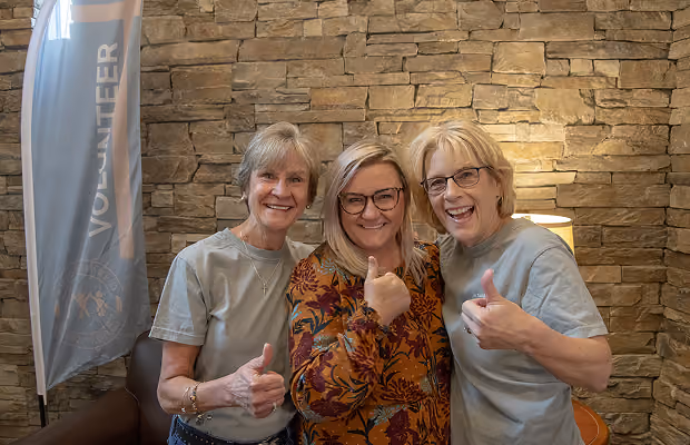 Three smiling women standing indoors in front of a stone wall, each giving a thumbs-up gesture; a vertical banner reading 'VOLUNTEER' is visible on the left.
