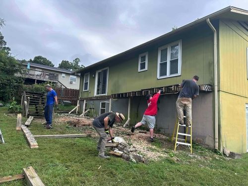 Four men working on house deck construction or renovation outside a green house with yellow base; one man stands on a ladder fixing the deck framing, another man in a red shirt supports the deck structure, a third man wearing a hat bends over picking up materials, and a fourth man in blue looks down near wooden planks and tools on the grass.