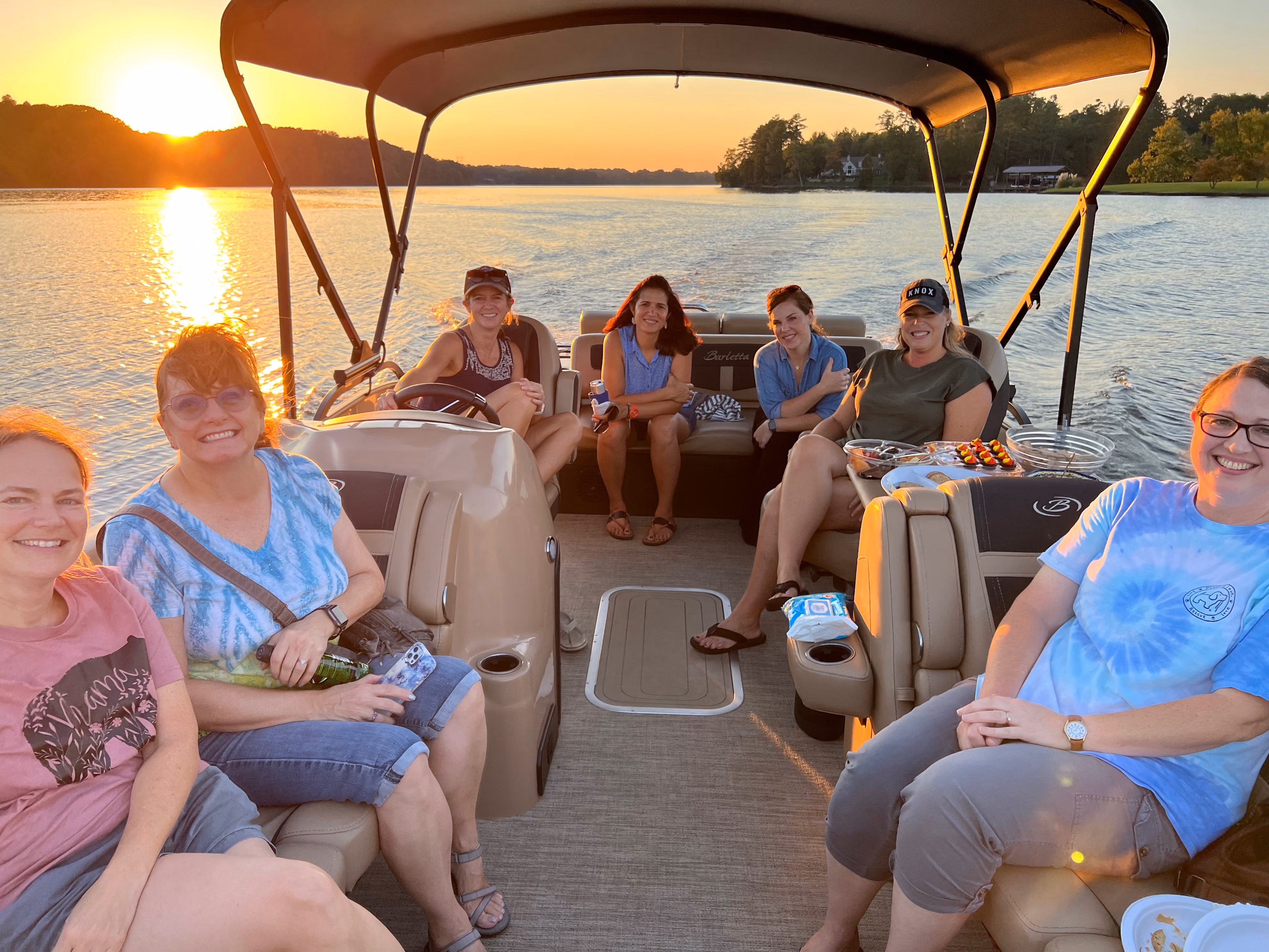 Seven women smiling and seated on a pontoon boat during sunset on a lake with trees and houses in the background, with snacks on a small table beside them.