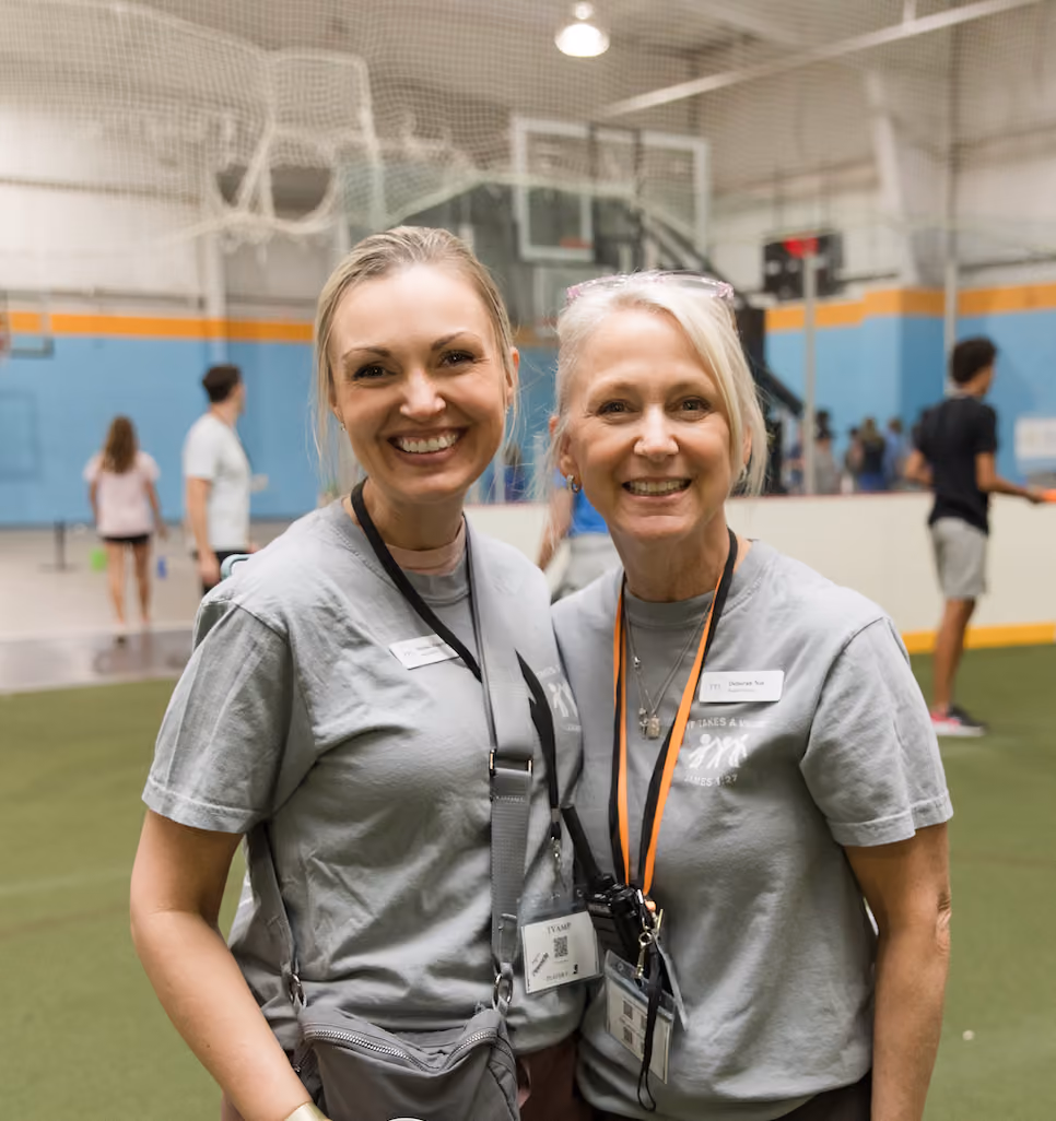 Two smiling women wearing gray T-shirts with name tags and lanyards, standing indoors on a sports court with blurred people in the background.