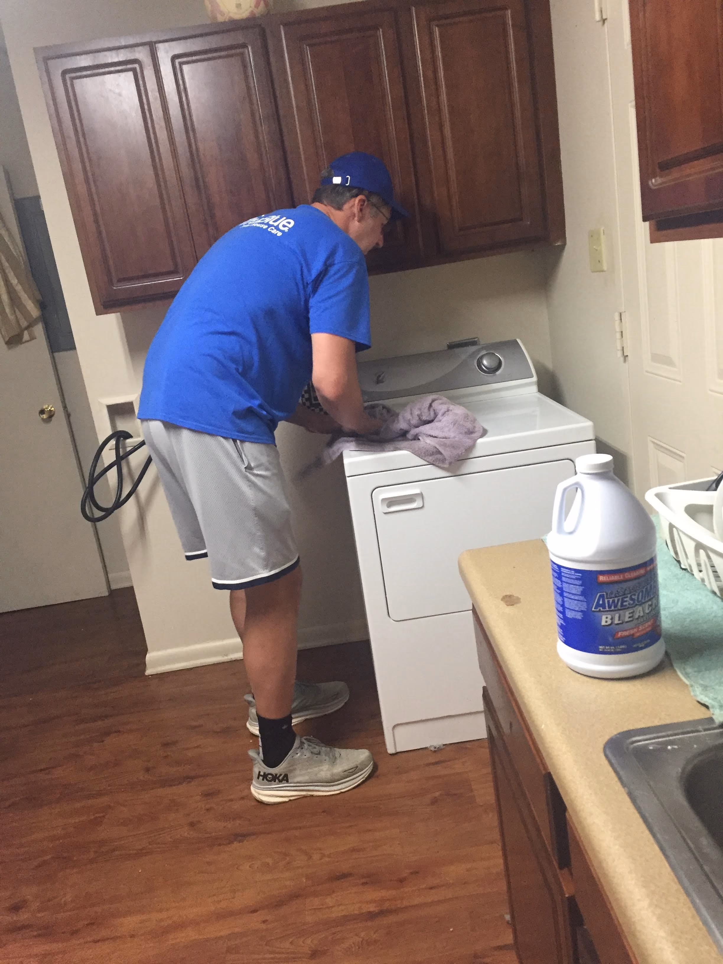 A man in a blue shirt, gray shorts, and white sneakers is loading or unloading a white clothes dryer in a home laundry room with wooden floors and cabinets.