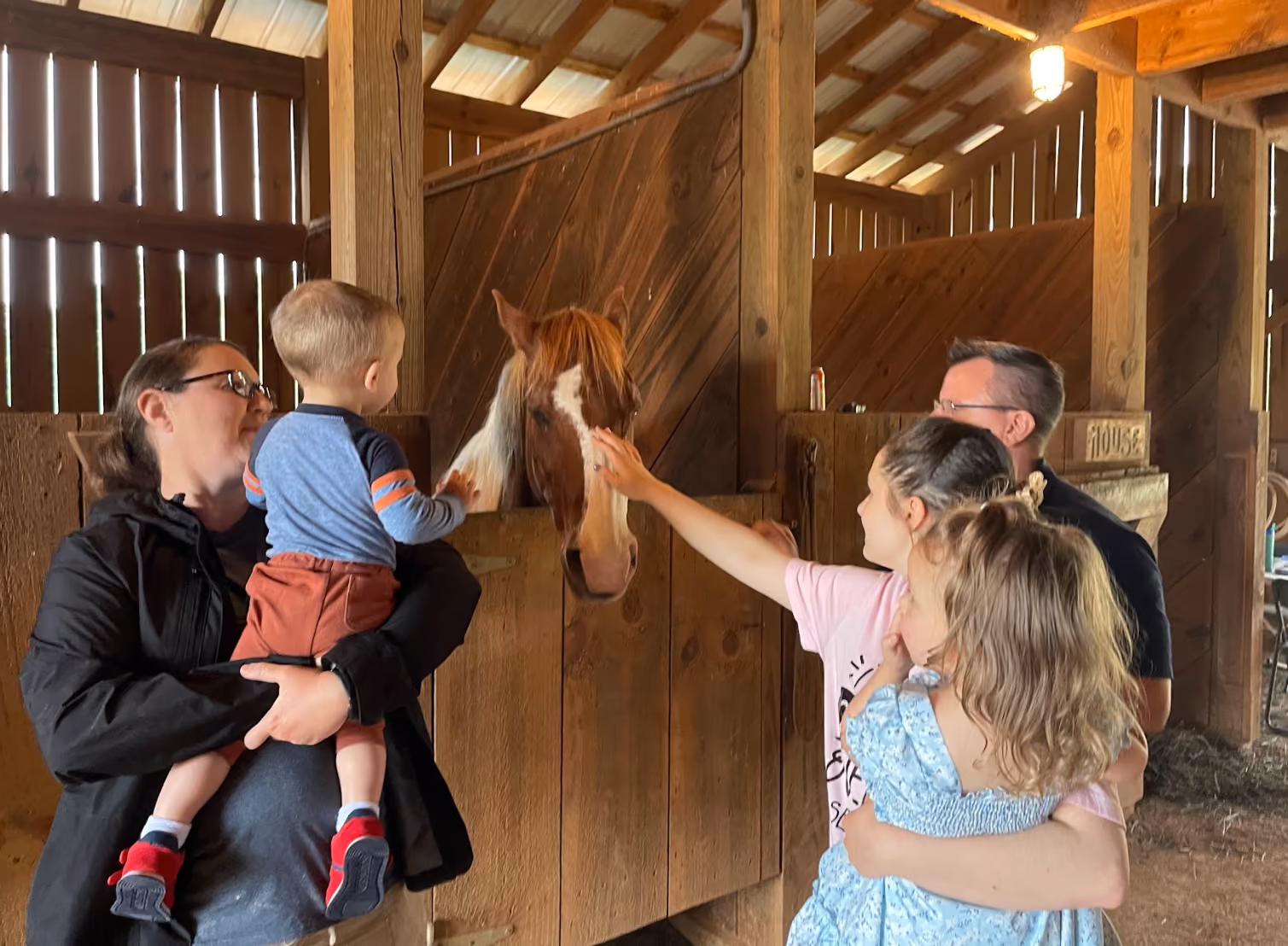 Two adults each holding a child are petting a brown and white horse inside a wooden stable with natural light coming through the slats.