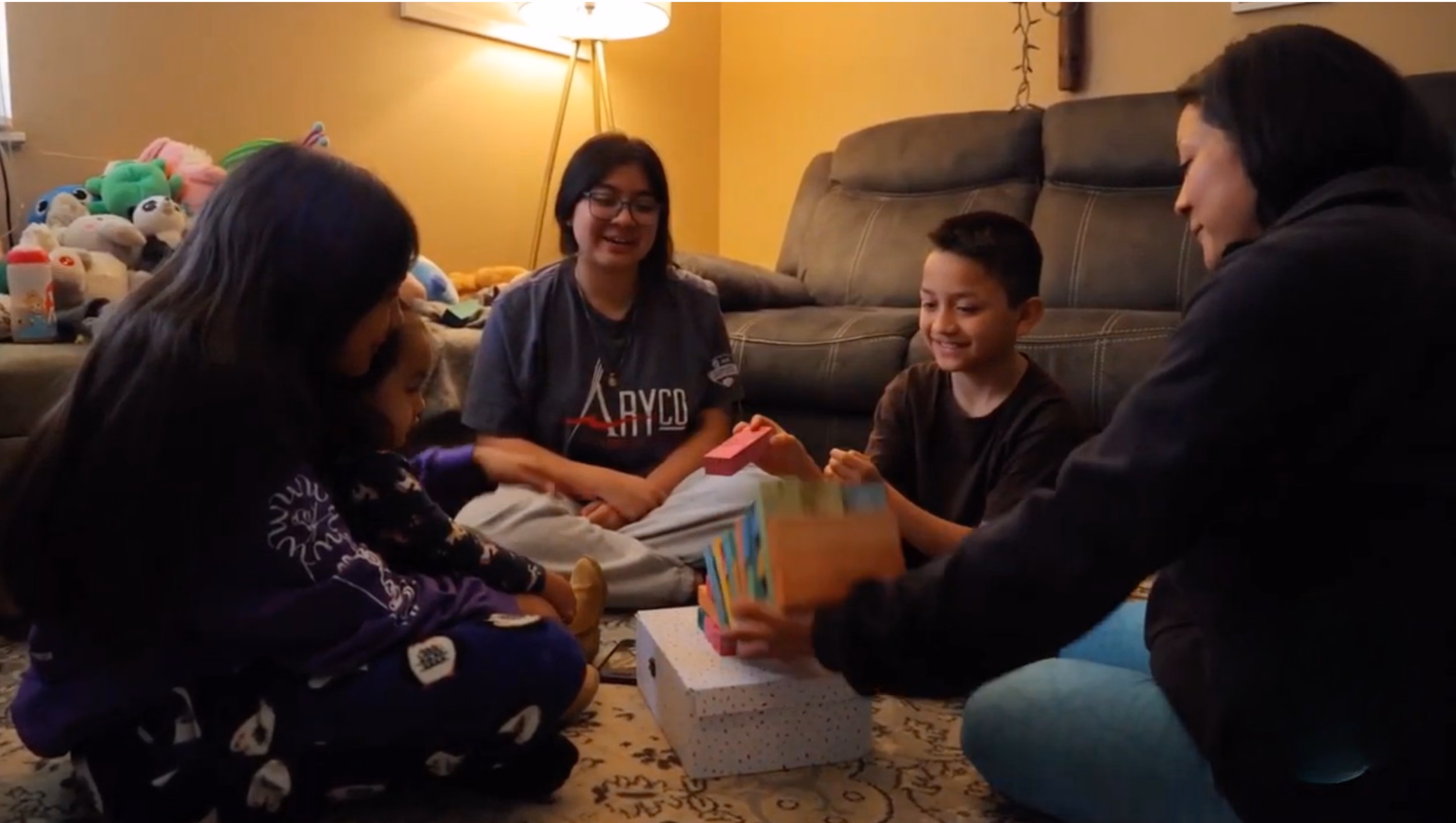 Four people, including a young boy stacking colorful blocks together on a white box in a living room while others watch and smile.