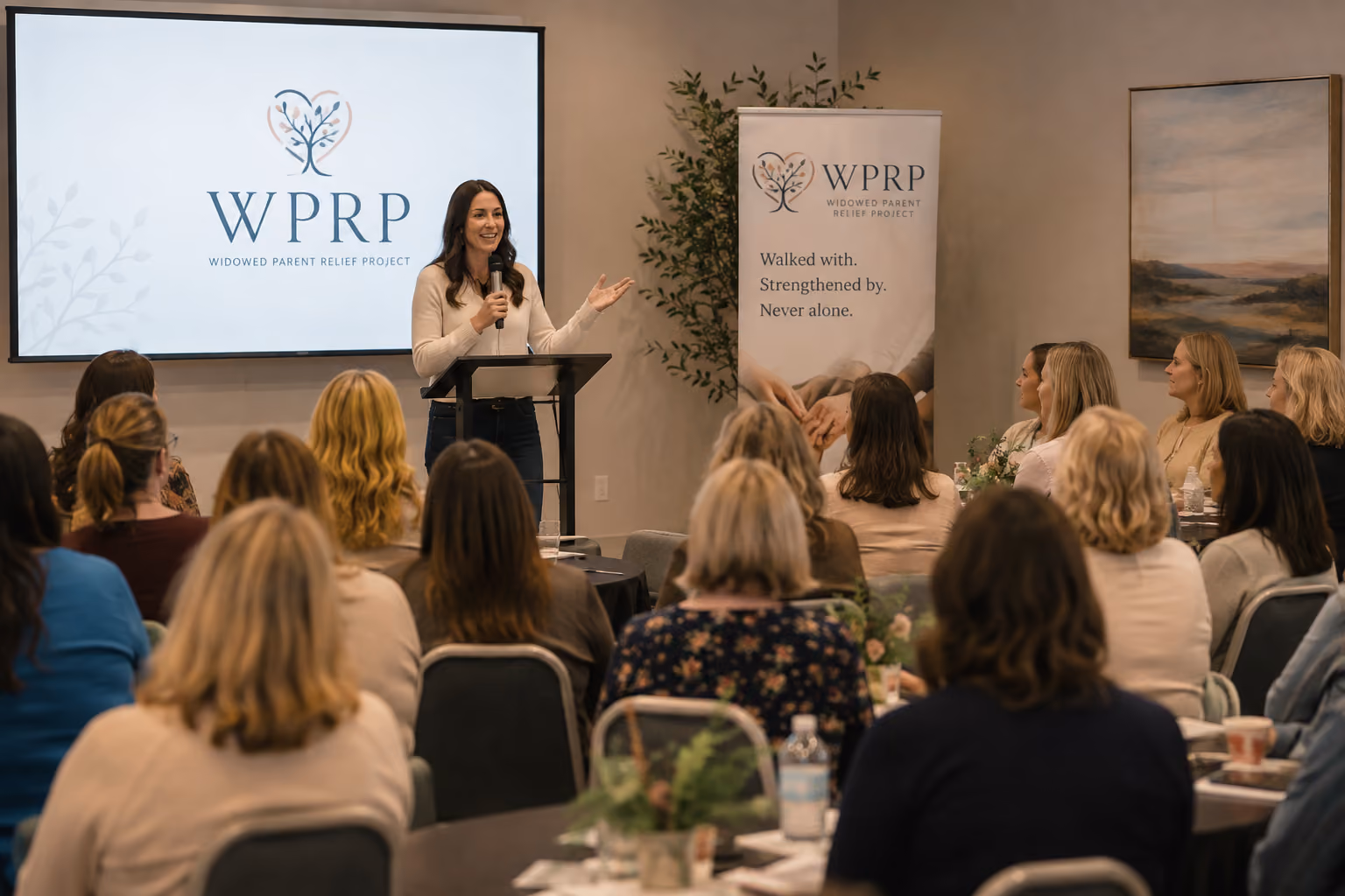 A woman stands at a podium speaking into a microphone to an audience of mostly women seated at tables in a conference room. Behind her is a screen showing the logo WPRP and the text Widowed Parent Relief Project. A banner to the side displays the same logo and the phrases: Walked with. Strengthened by. Never alone.
