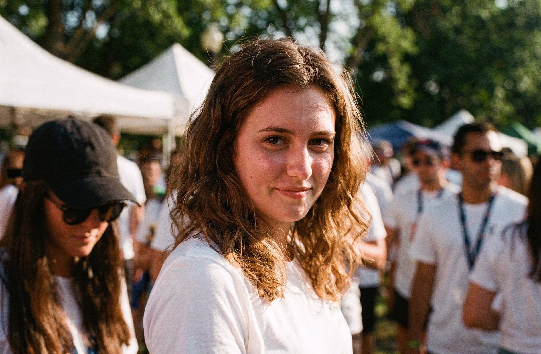 Young woman with curly brown hair wearing a white shirt looking at the camera at an outdoor event with people and tents in the background.