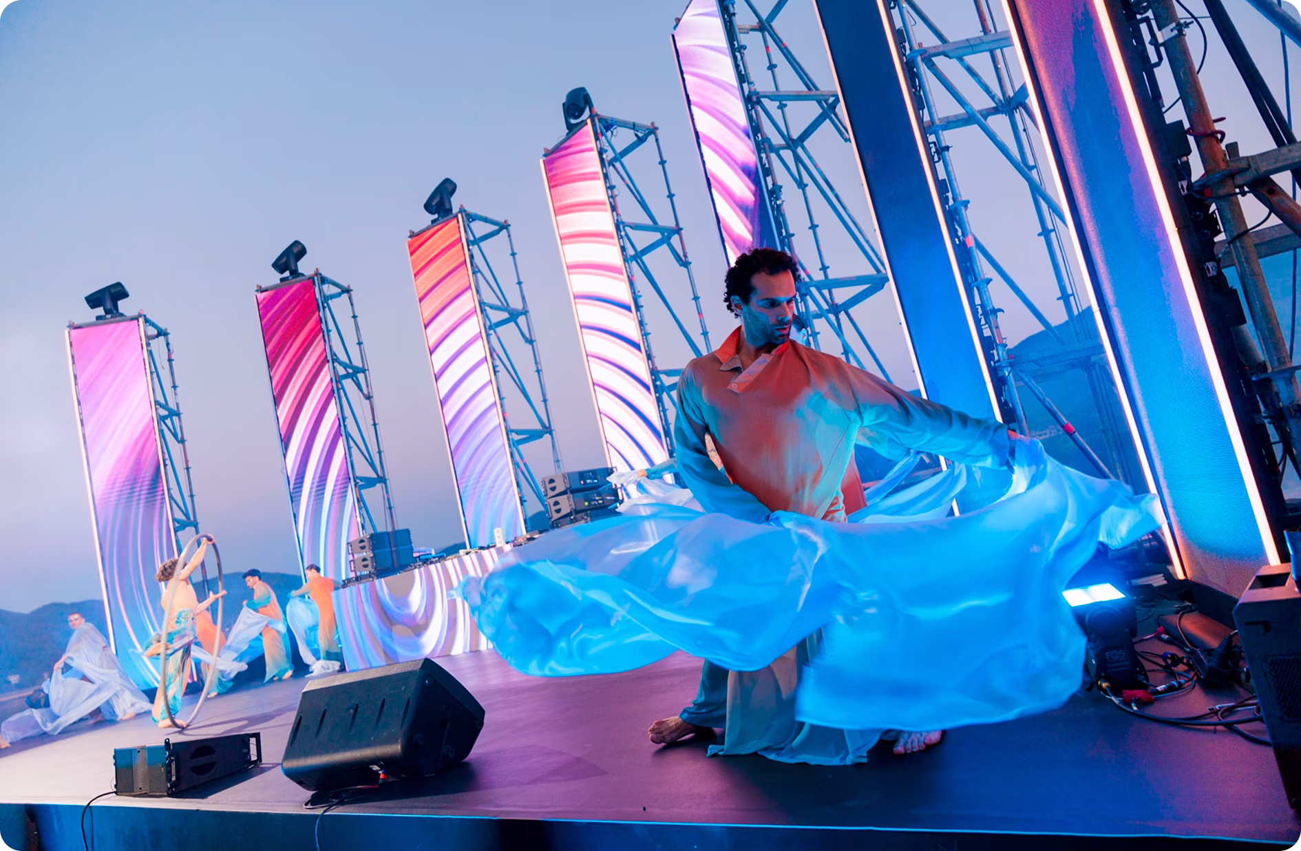 Male dancer in flowing blue costume performing on stage with colorful vertical LED panels and other performers in the background.