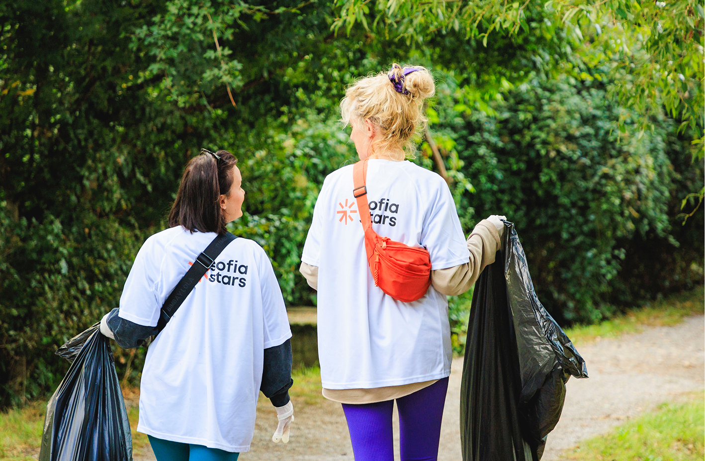 Two smiling women wearing sunglasses stand close together by a pool, one in a floral dress and the other in a white t-shirt and beige pants.