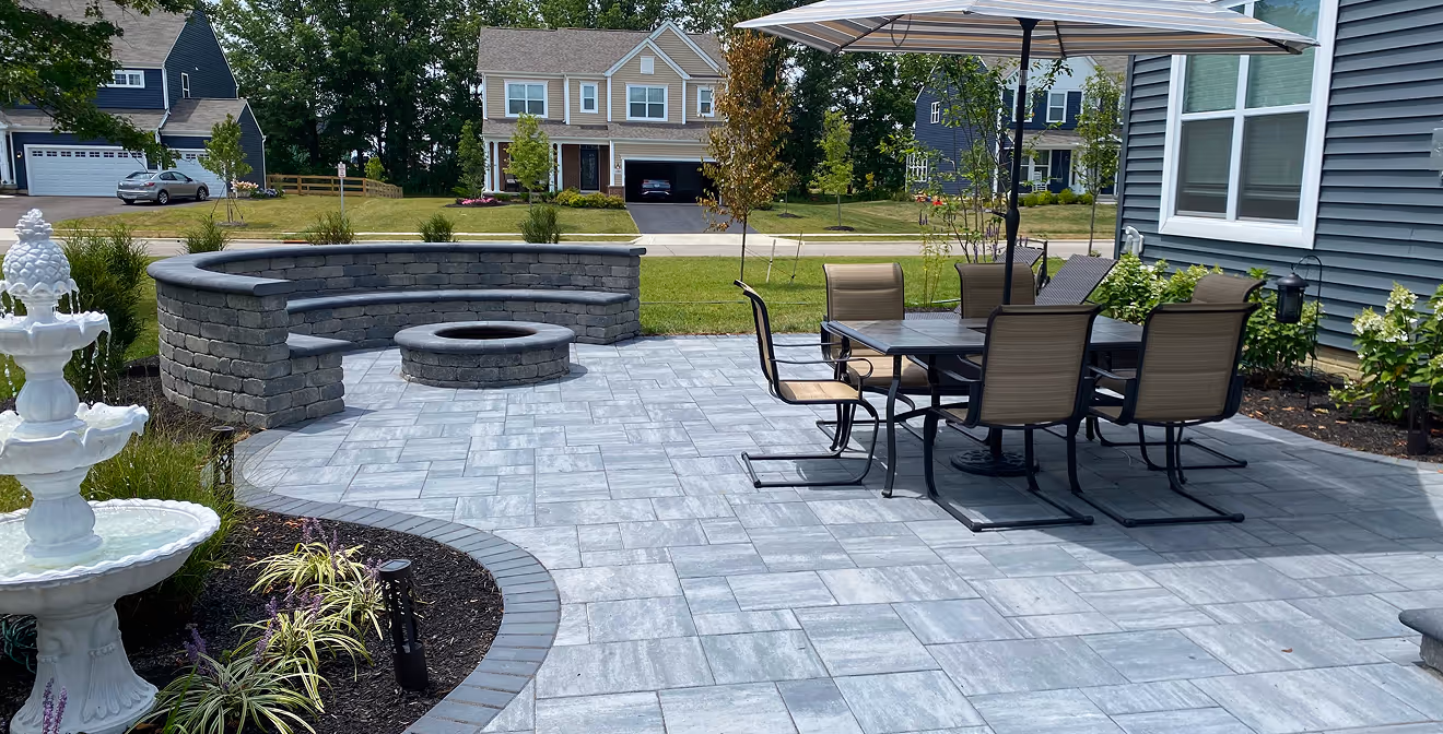 Backyard patio with gray patterned stone tiles, a curved stone bench surrounding a fire pit, and a dining table with six chairs under a large umbrella.