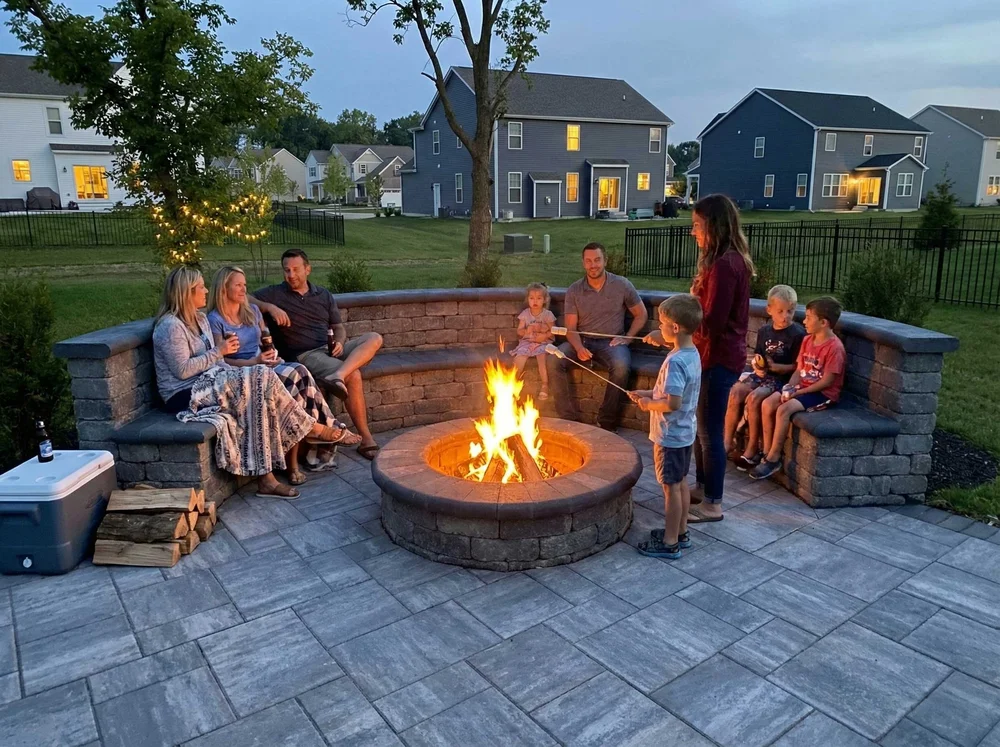 Family and friends gathered around a lit fire pit in a backyard patio, roasting marshmallows in the evening.