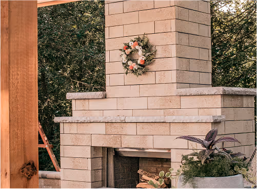 Outdoor brick fireplace decorated with a floral wreath and surrounded by greenery and potted plants.