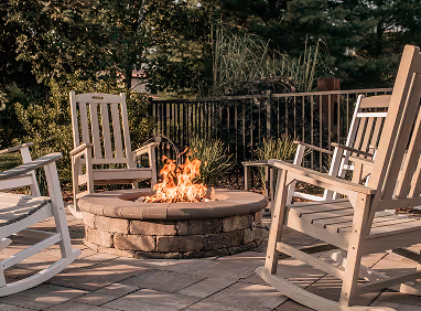 Outdoor stone fire pit surrounded by four wooden rocking chairs on a stone patio with green trees in the background.