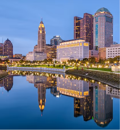 City skyline at dusk with illuminated buildings reflected in a calm river.