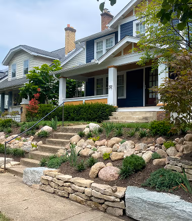 Two-story house with blue siding, white columns on front porch, stone steps, and landscaped garden with rocks and shrubs.