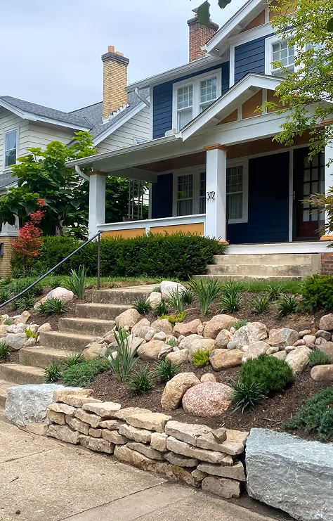 Blue house with white trim, front porch, stone retaining wall, and landscaped garden with rocks and plants.