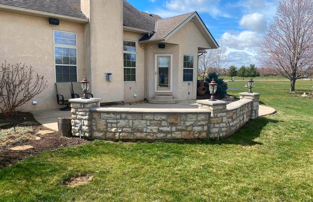 Stone patio with decorative lamps in front of beige house with large windows, surrounded by green lawn and trees.