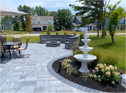 Patio with gray stone tiles, a black metal dining table with chairs under an umbrella, a curved stone fire pit wall, and a tiered white garden fountain surrounded by plants.