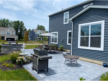 Backyard patio with gray stone tiles, a barbecue grill, outdoor dining table with umbrella, and a water fountain near a blue-gray house.