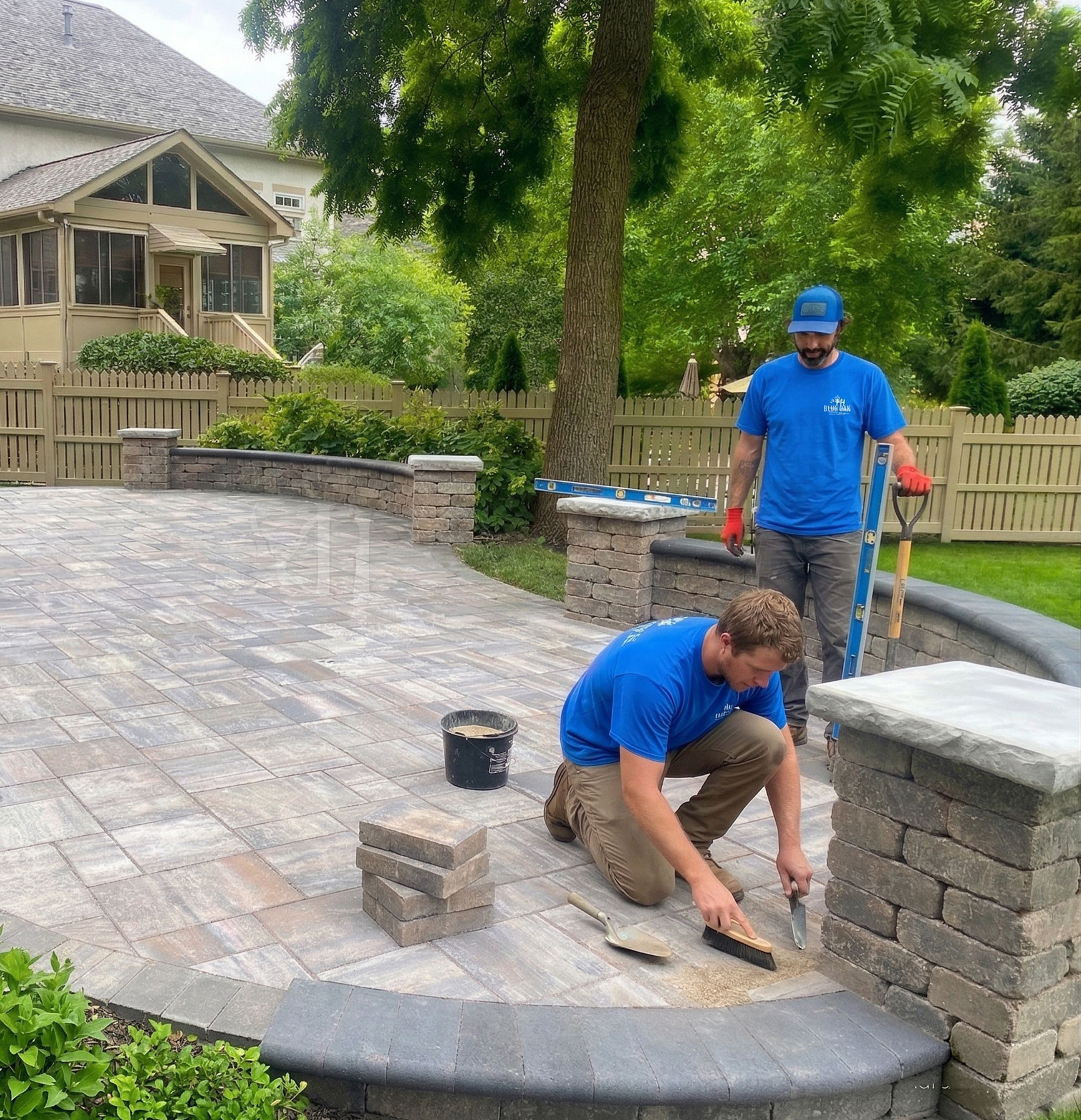 Two men in blue shirts working on installing stone pavers in a backyard patio area with green trees and a beige fence in the background.