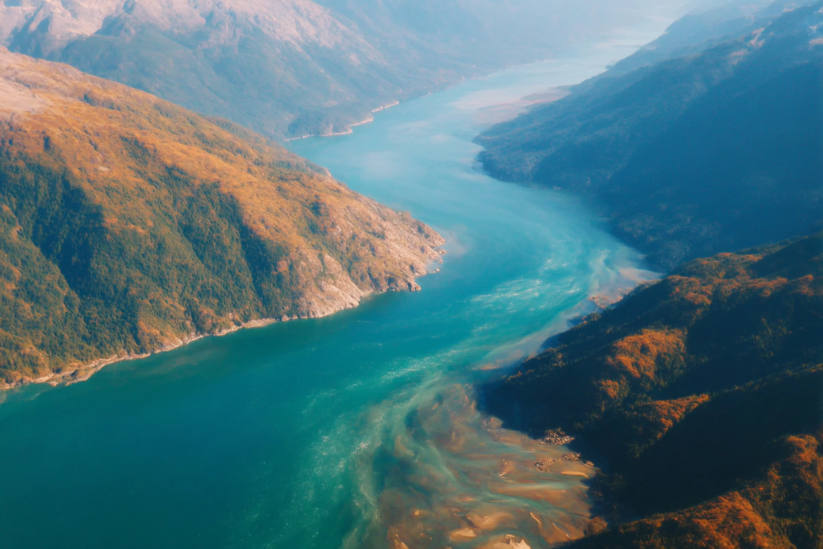 Aerial view of a winding fjord surrounded by steep, forested mountains with autumn foliage.