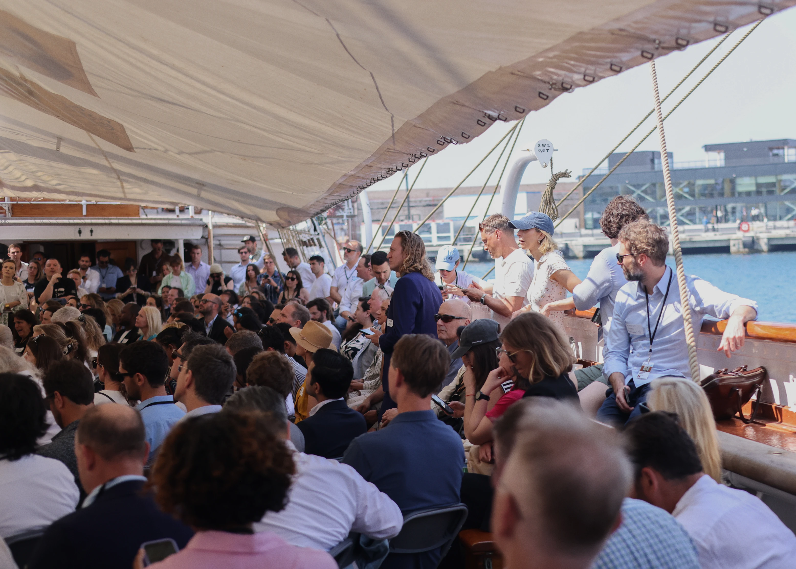 Large group of people seated on a covered outdoor boat deck near a waterfront, engaged in conversation and using mobile devices.