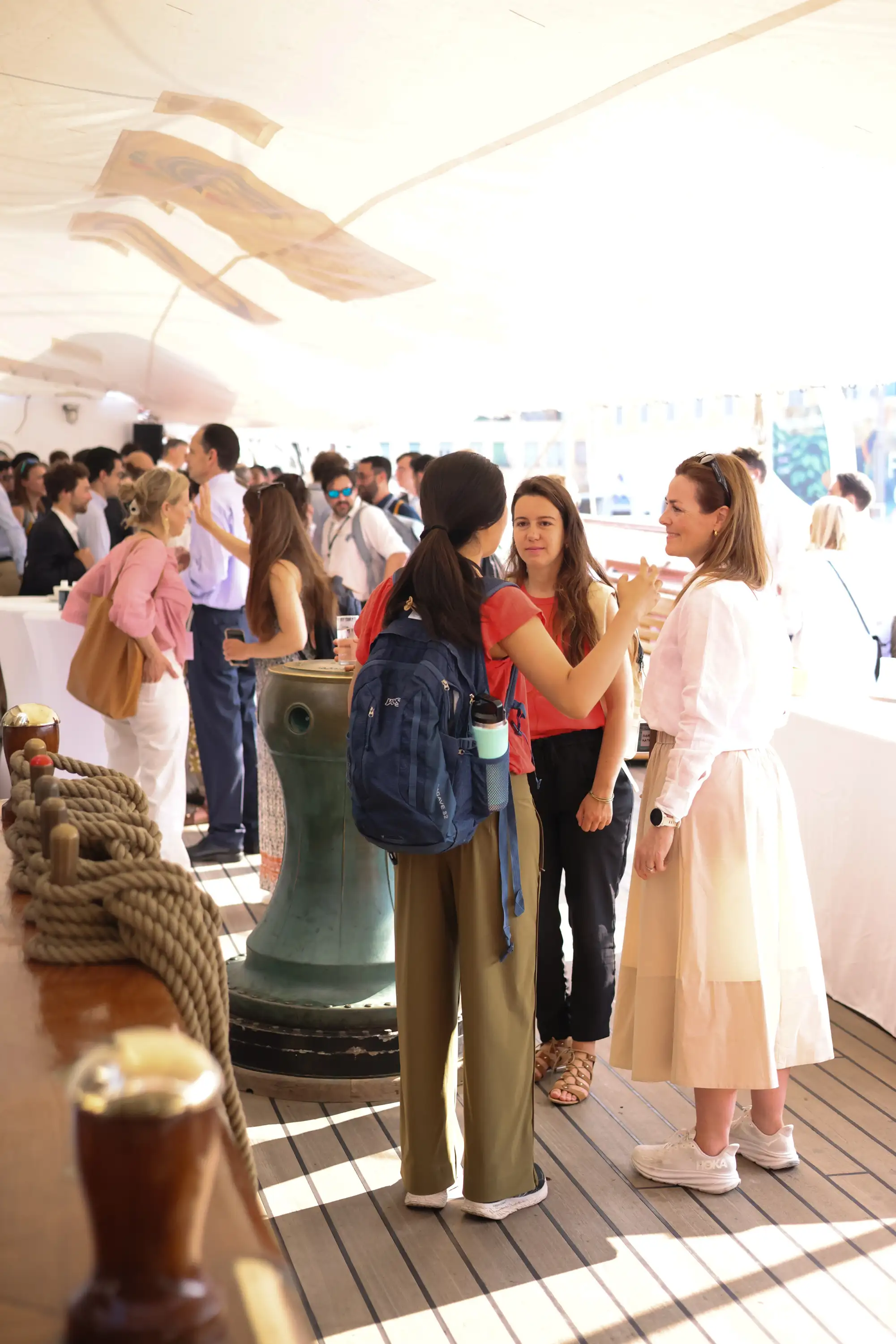 Group of people mingling and chatting casually under a tent with wooden flooring, some holding drinks.