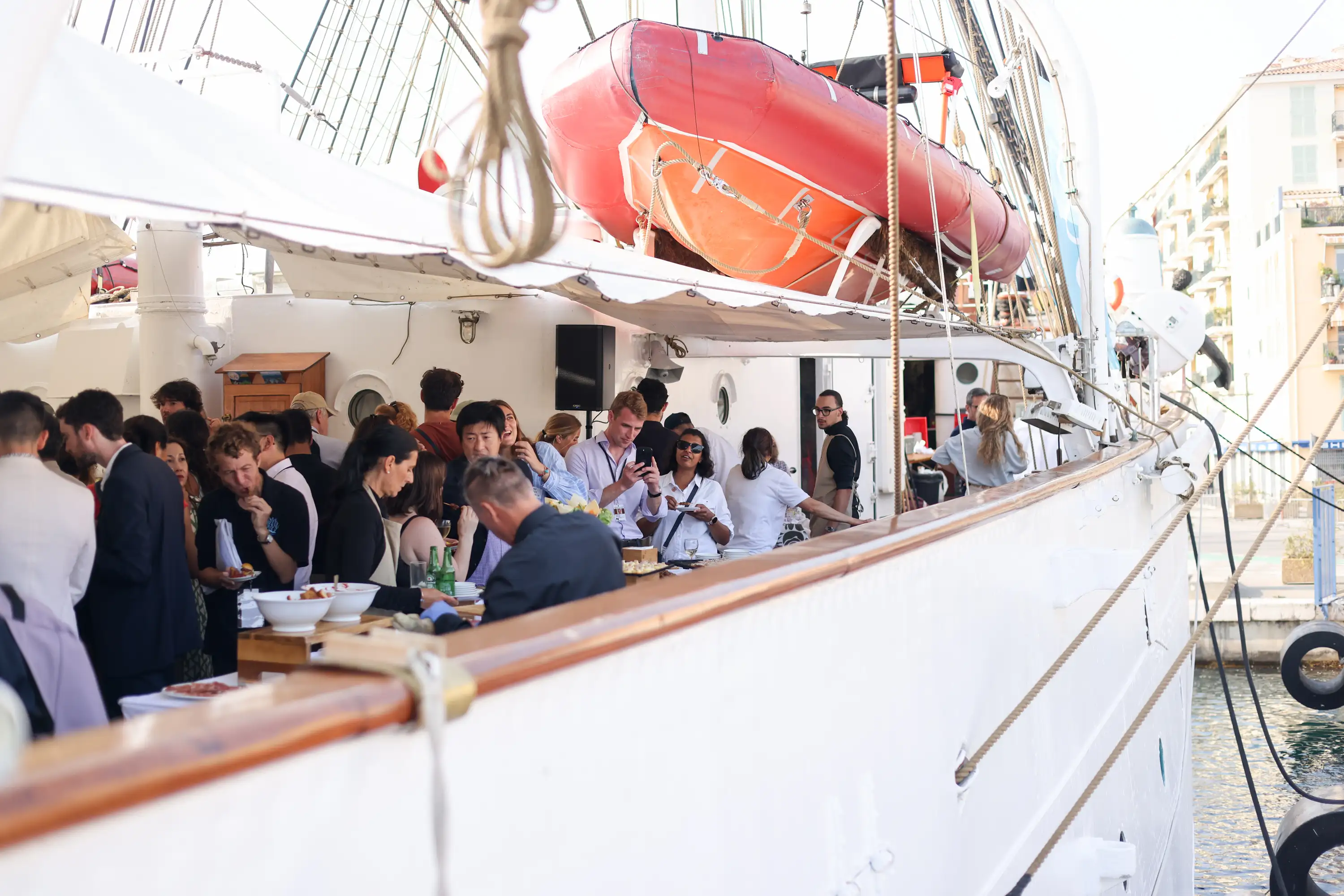 People socializing and enjoying food on the deck of a docked white ship with a red lifeboat overhead.