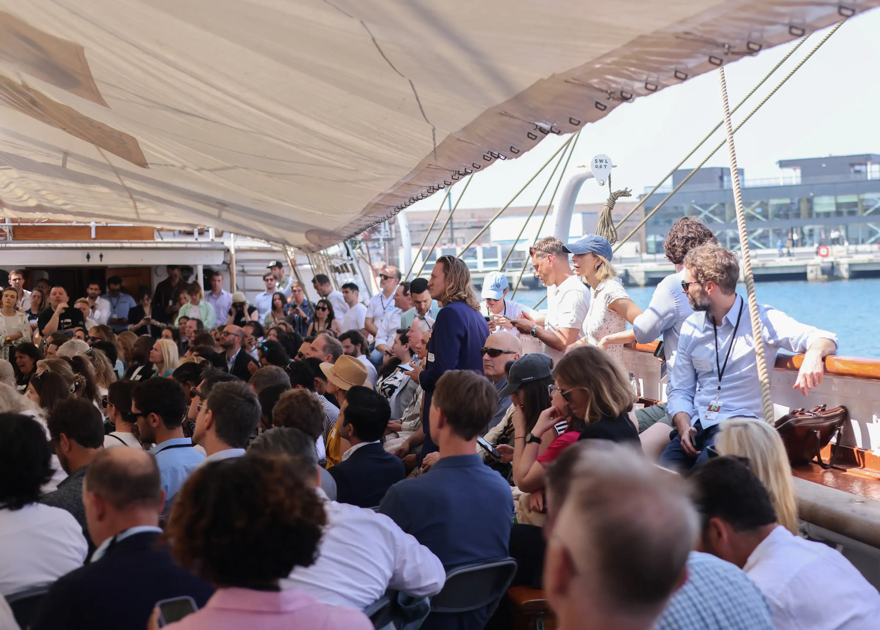 Crowd of people seated under sail shade on a boat near a harbor, some using phones or talking.
