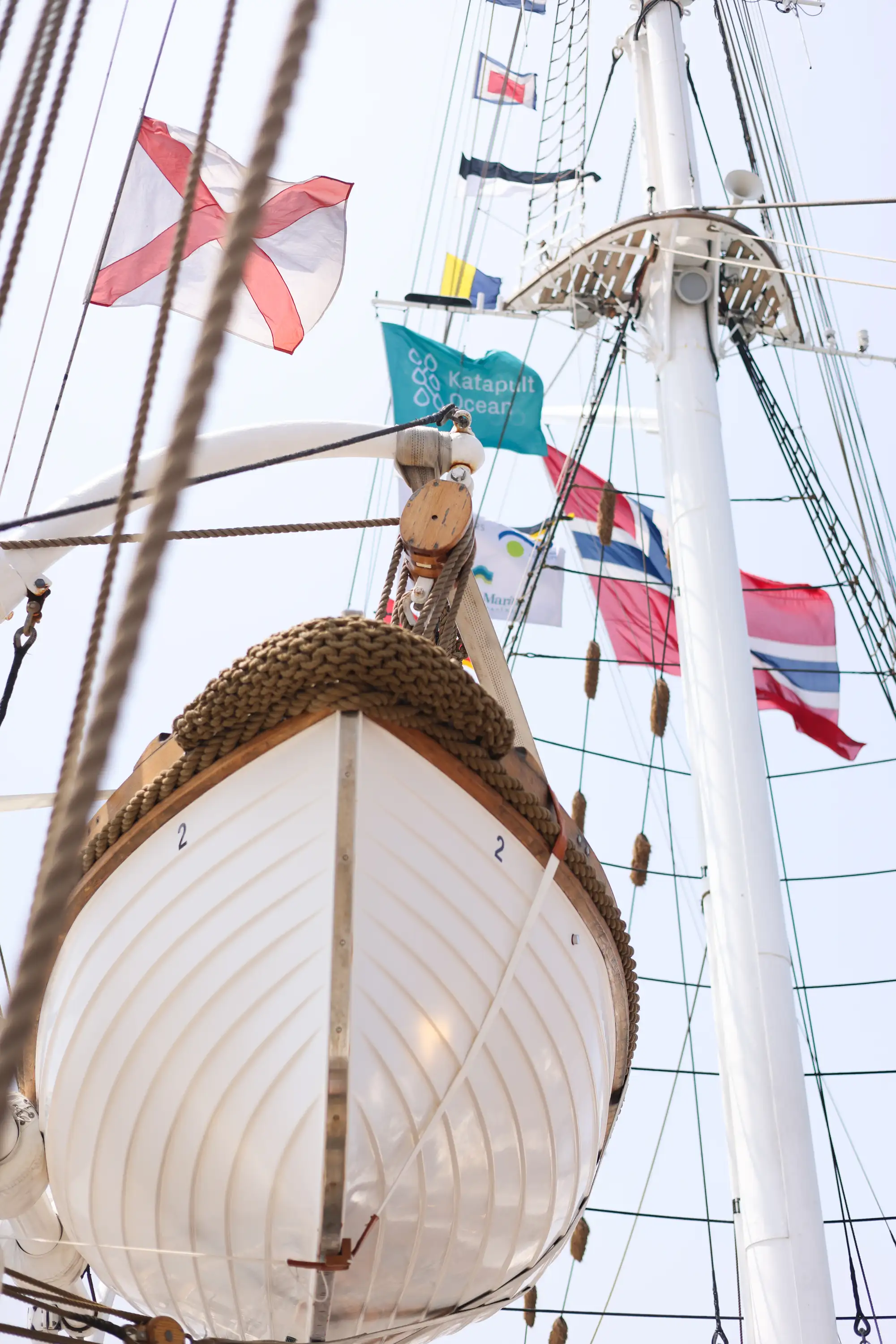 White lifeboat numbered '2' hanging on the side of a tall ship with rigging and multiple flags in the background.