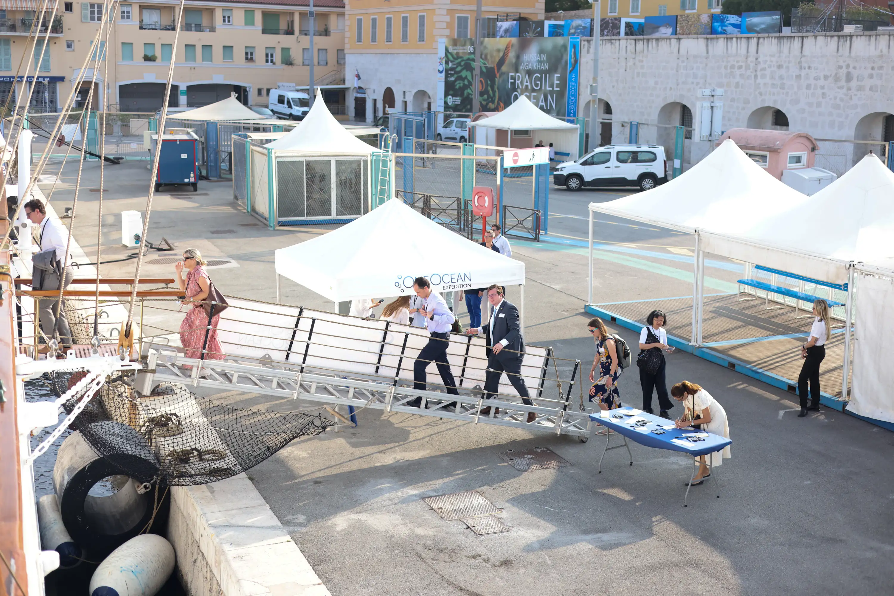 People boarding a ship via a gangway at a dock with white tents and buildings in the background.