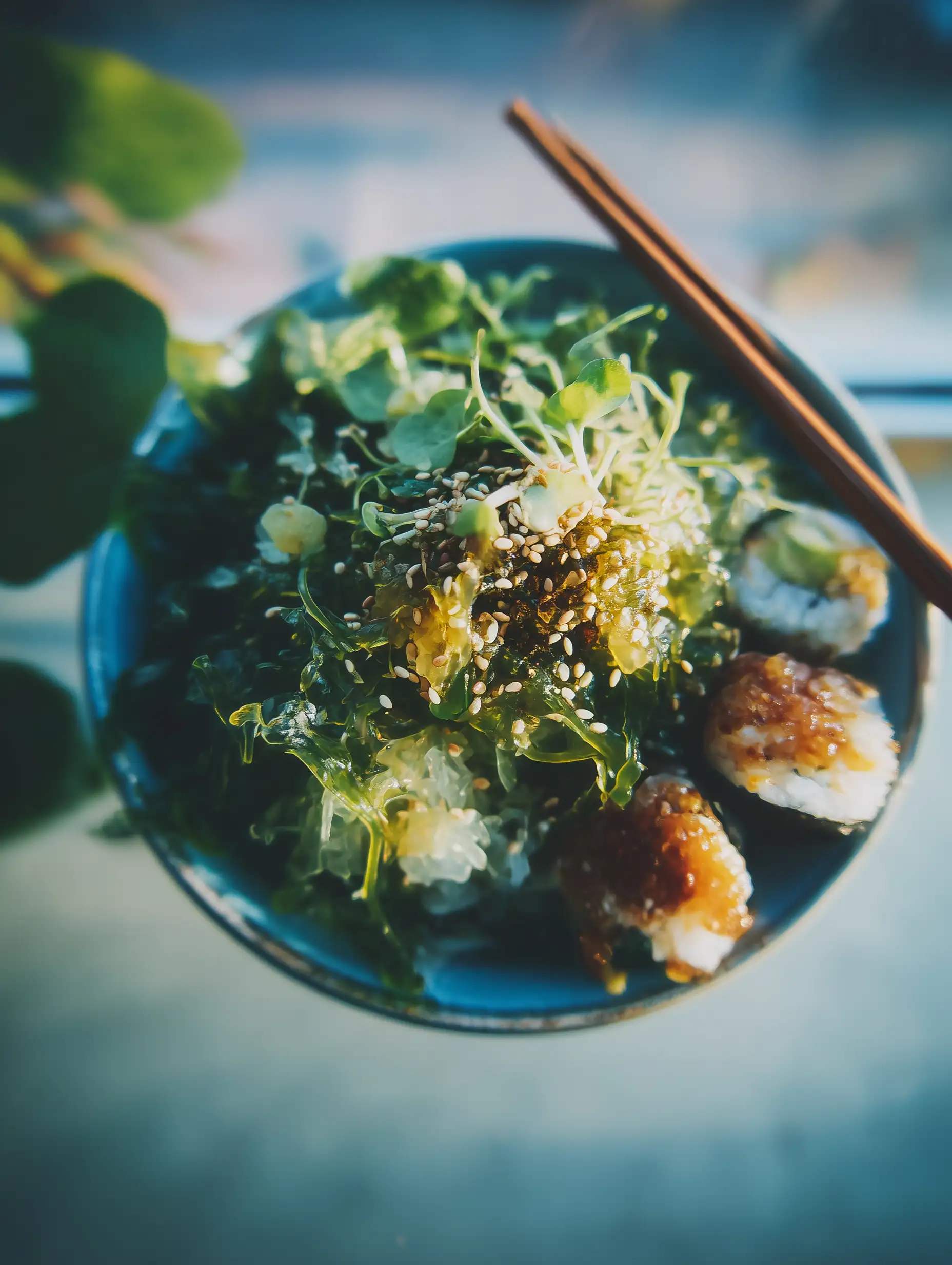 Bowl of seaweed salad topped with sesame seeds and microgreens, with pieces of sushi and chopsticks on the side.