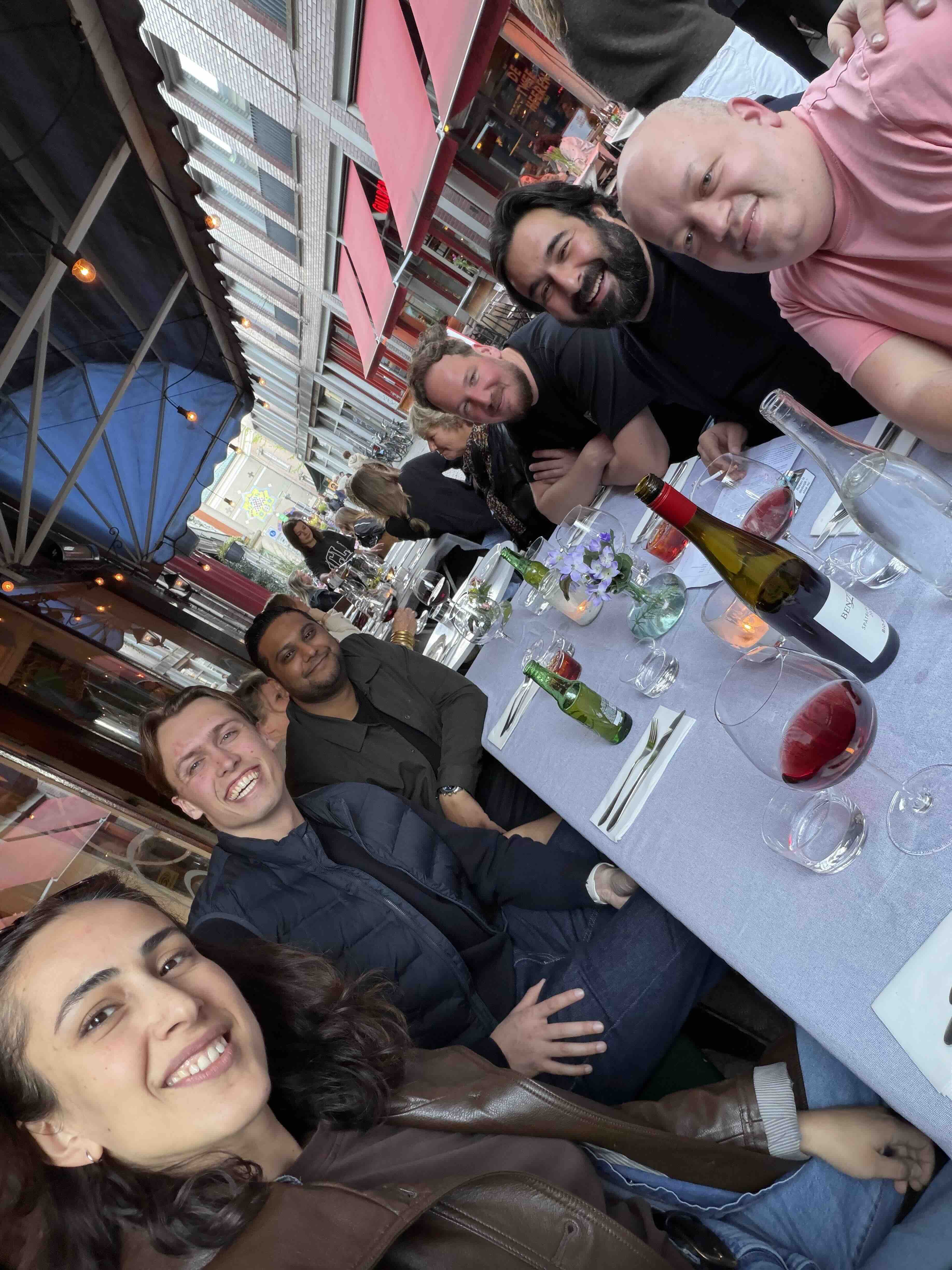 Vijf vrienden zitten aan een lange buitentafel met een lichtblauw tafelkleed, glazen wijn, bierflessen en een fles water, lachend naar de camera.