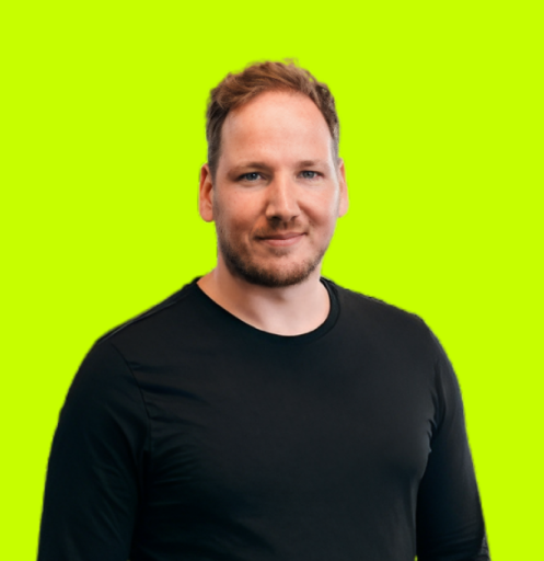 Man met blond haar en baard, gekleed in een zwarte trui, staand voor een groene achtergrond.