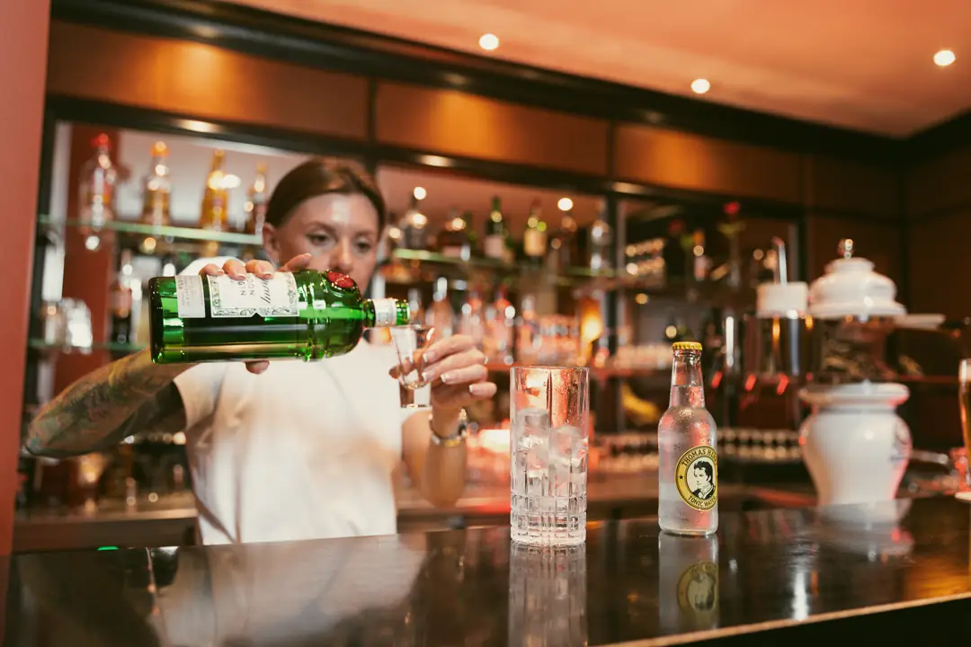 Barkeeperin gießt einen klaren Schnaps in ein Glas in einer Bar mit Flaschenregal im Hintergrund.