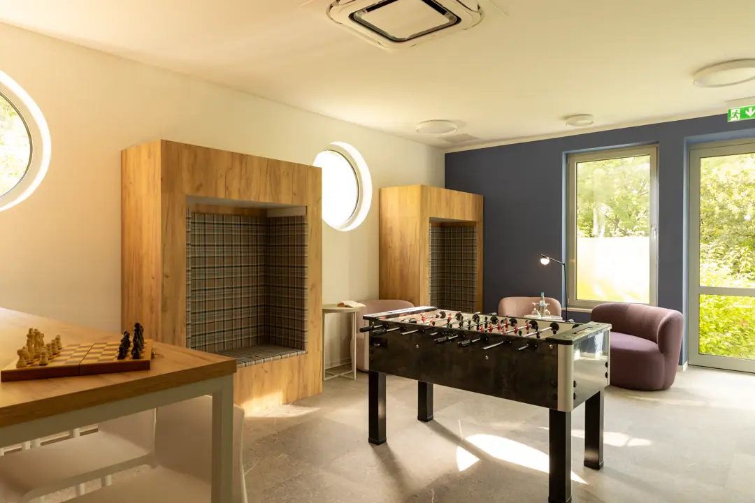 Gemütlicher Aufenthaltsraum mit einem Kicker-Tisch, einem Schachspiel auf einem Holztisch und zwei Sitznischen mit karierten Polstern.