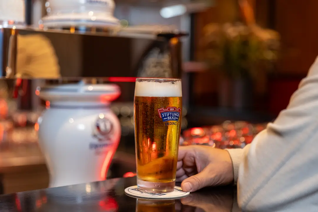 Ein Glas Bier der Marke Stiftungsbräu mit Schaum steht auf einem Bierdeckel an einer Bar, daneben liegt eine Hand.