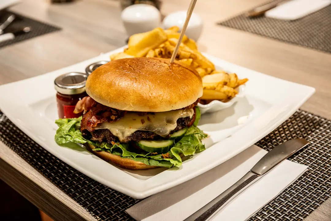 Burger mit Speck, Käse, Salat und Gurken auf einem weißen Teller mit Pommes und zwei kleinen Gläsern mit Soße.
