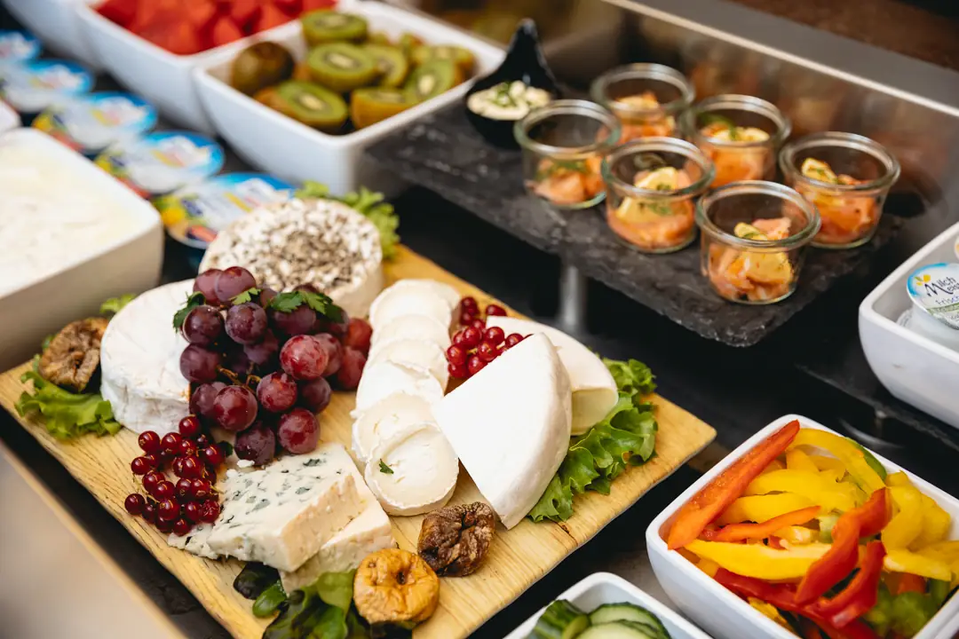 Vielfältige Käseplatte mit Trauben, roten Johannisbeeren, getrockneten Feigen und Salat, umgeben von frischem Obst, Gemüse und kleinen Gläsern mit Lachs.