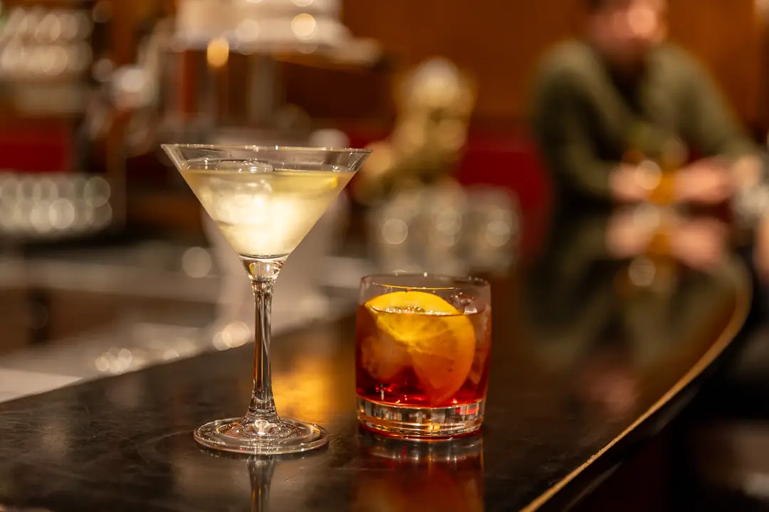 Zwei Cocktails auf einer Theke, ein Martini-Glas mit Eis und ein Glas mit orangefarbener Scheibe.