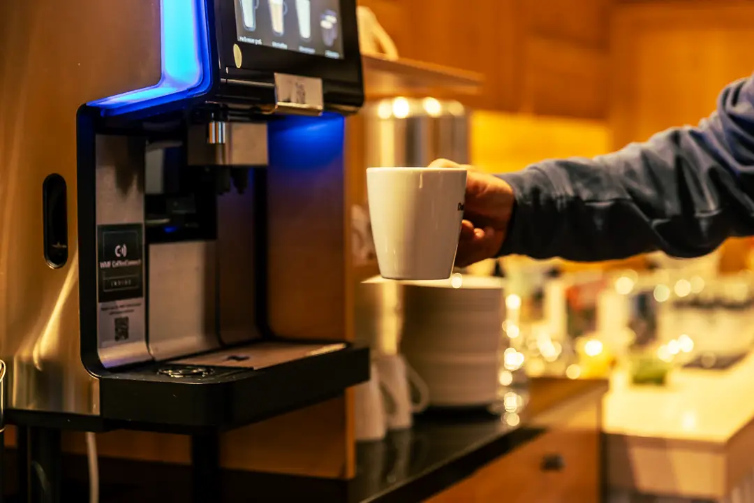 Person hält eine weiße Tasse unter einem Kaffeevollautomaten in einem warm beleuchteten Raum.