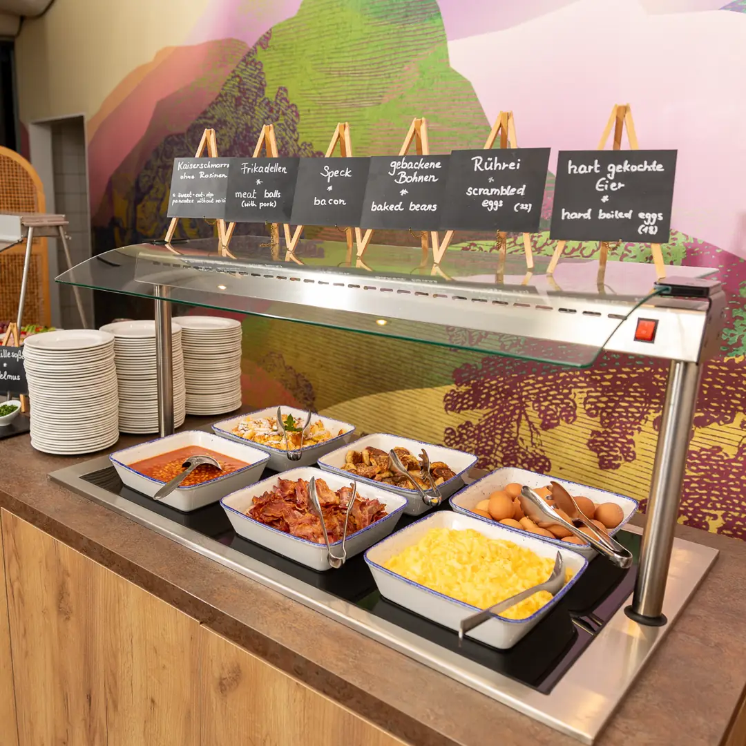 Frühstücksbuffet mit Tellern und warmen Speisen wie Rührei, hart gekochte Eier, Speck, gebackene Bohnen, Frikadellen und Kaiserschmarrn.