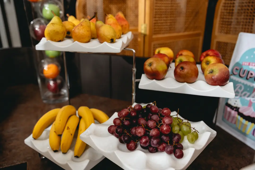 Mehrstöckiges Obstregal mit Birnen oben links, Äpfeln oben rechts, Bananen unten links und roten sowie grünen Trauben unten rechts.