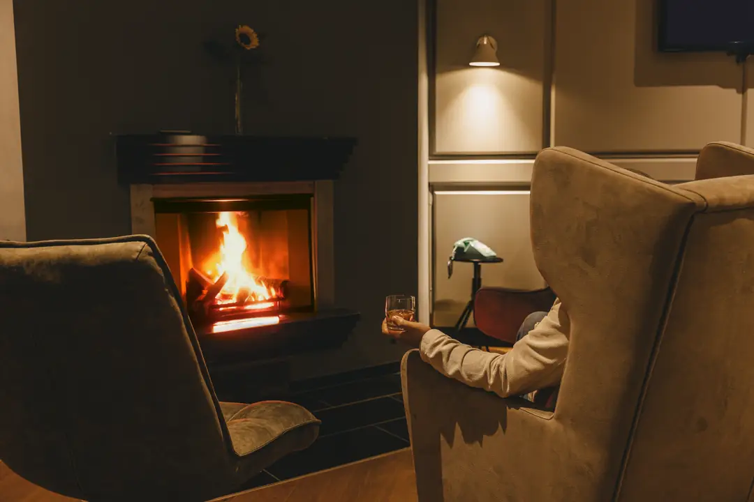Person sitzt in einem gemütlichen Sessel vor einem Kaminfeuer und hält ein Glas Whisky in der Hand.