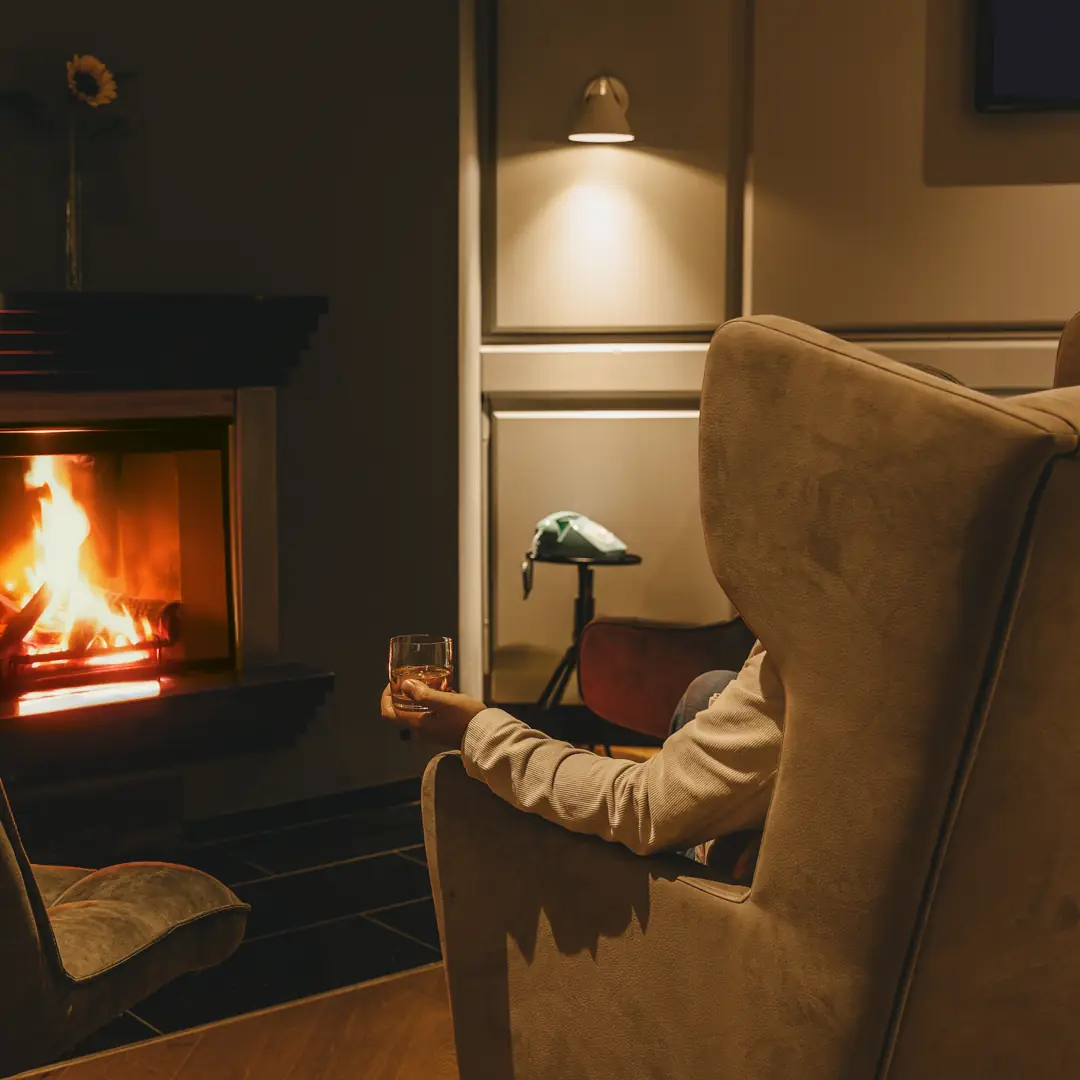 Person sitzt in einem gemütlichen Sessel vor einem Kamin und hält ein Glas Whisky in der Hand.