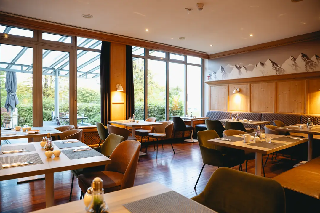 Hell eingerichtetes Restaurant mit Holztischen, gepolsterten Stühlen und großen Fenstern mit Blick ins Grüne.