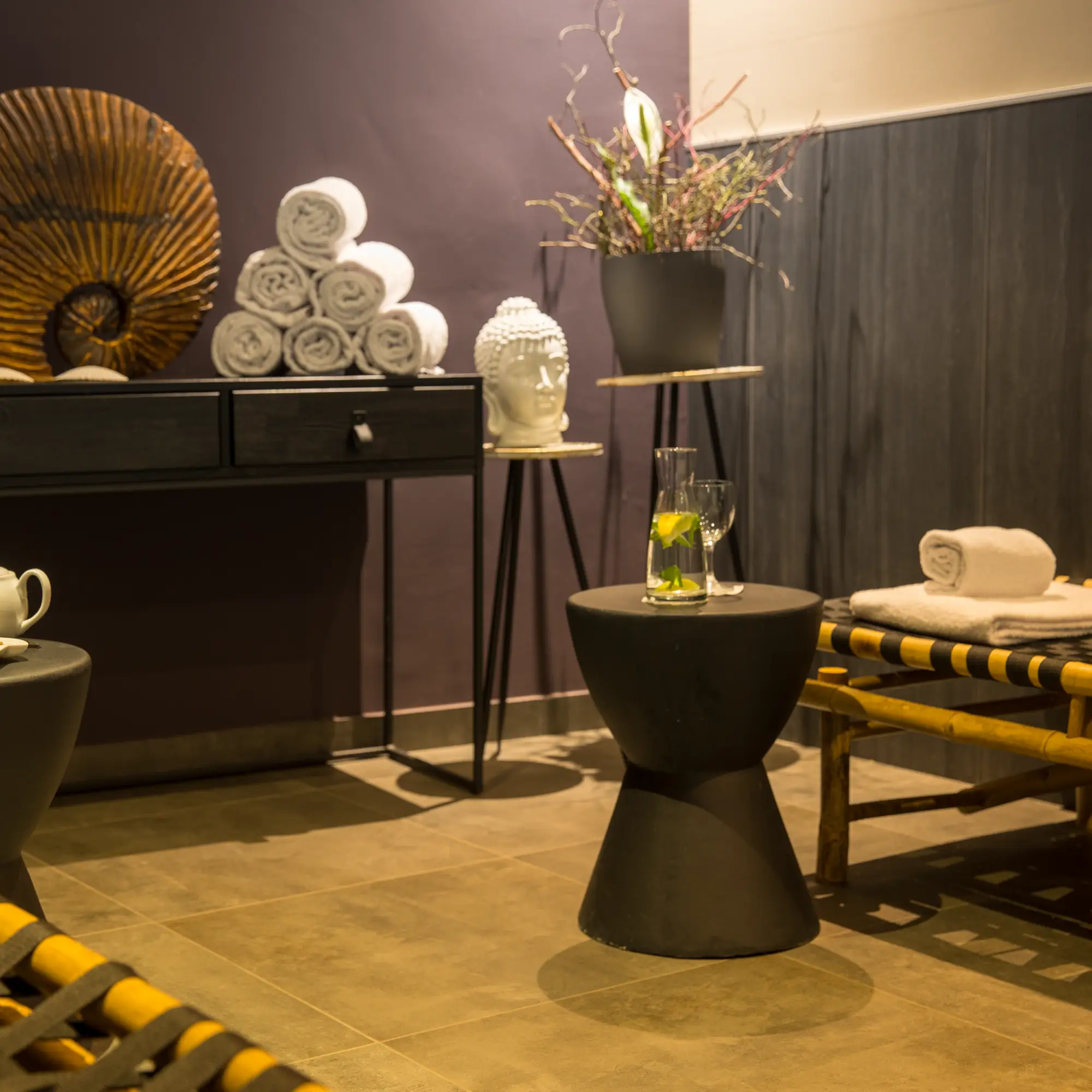 Entspannungsraum mit zusammengerollten Handtüchern, Buddha-Skulptur, Vase mit Zweigen und Wasserkrug mit Glas auf einem schwarzen Beistelltisch.