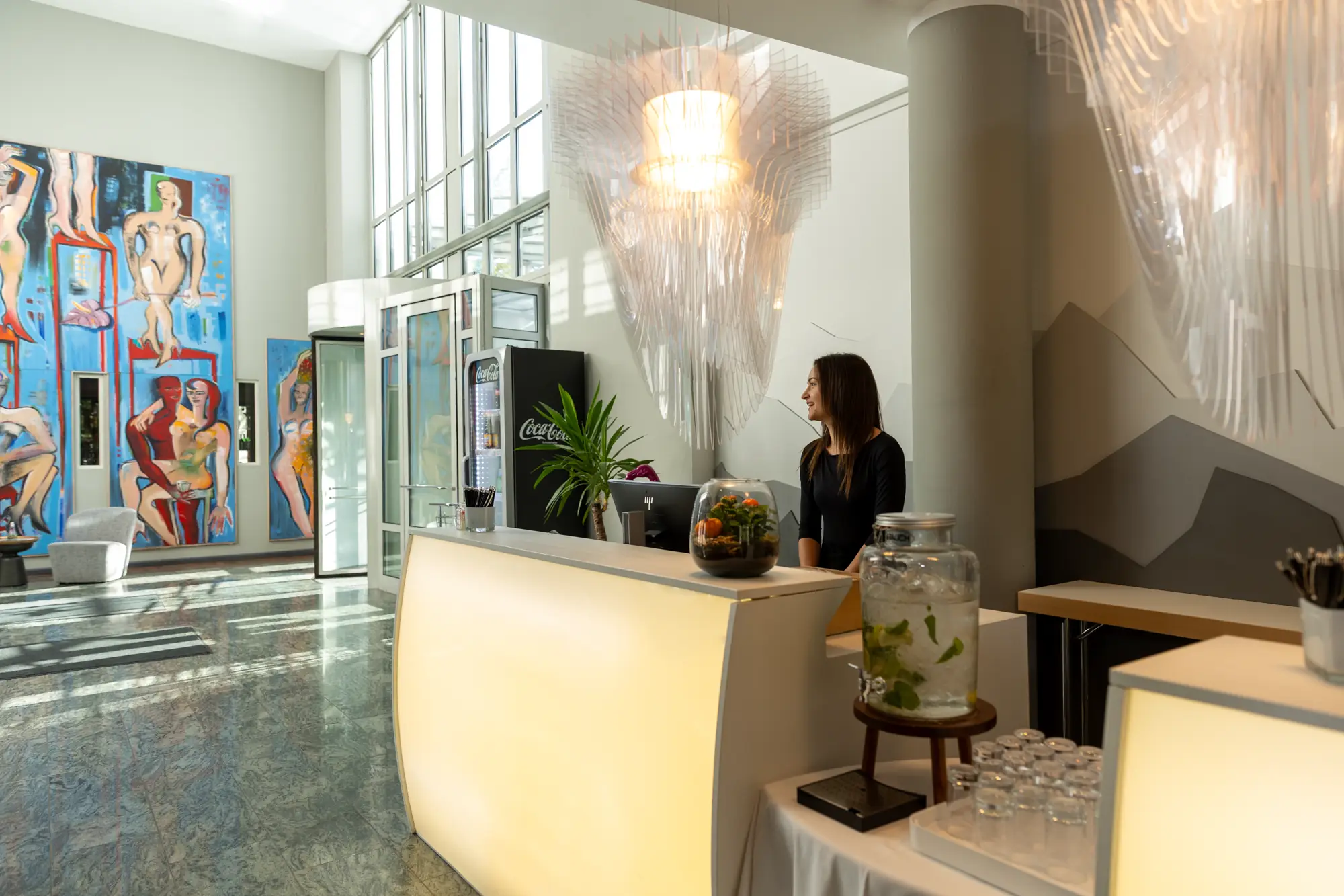 Moderne Hotelrezeption mit beleuchtetem Empfangstresen, einer Frau am Empfang, Wasserstation und farbenfrohen Kunstwerken an der Wand.