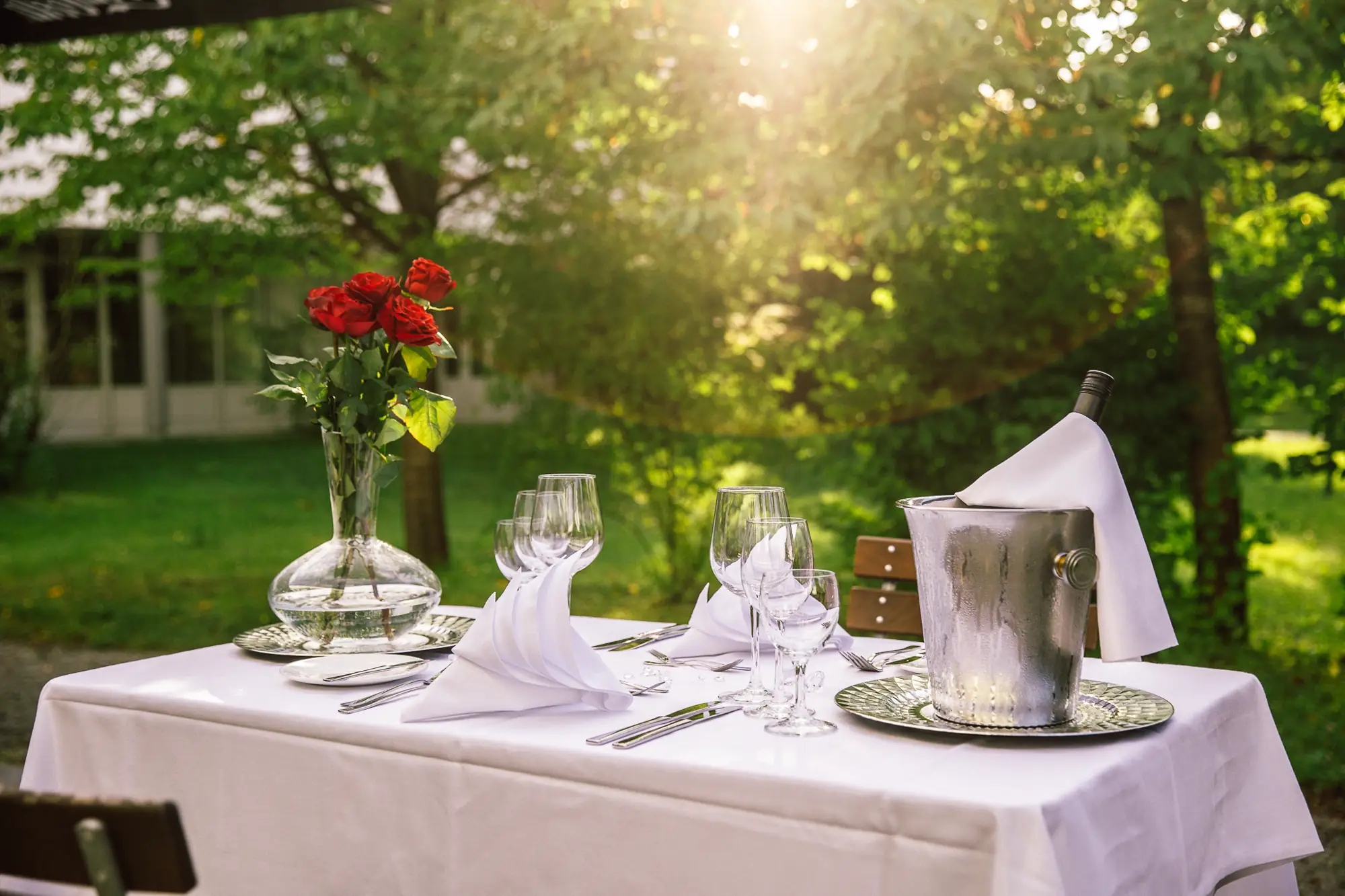Romantisch gedeckter Tisch im Freien mit weißen Servietten, Weingläsern, Rosenstrauß und Weinflasche im Eisbehälter.