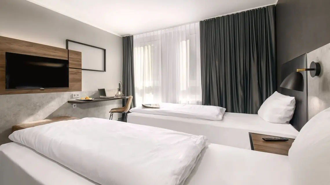 Modernes Hotelzimmer mit zwei Einzelbetten, Fernseher an der Wand, Schreibtisch mit Stuhl und großen Fenstern mit Vorhängen.